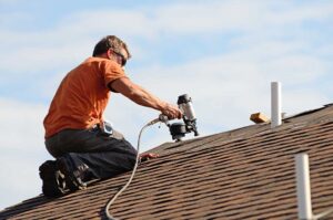 A raccoon clambers near the edge of a residential roof, inspecting loose shingles and a vent cover.